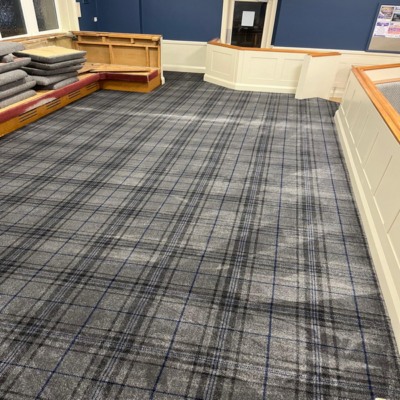 A room with blue walls and a plaid-patterned carpet. Stacked carpet pieces are visible in the corner. White paneling and a wooden railing are present.