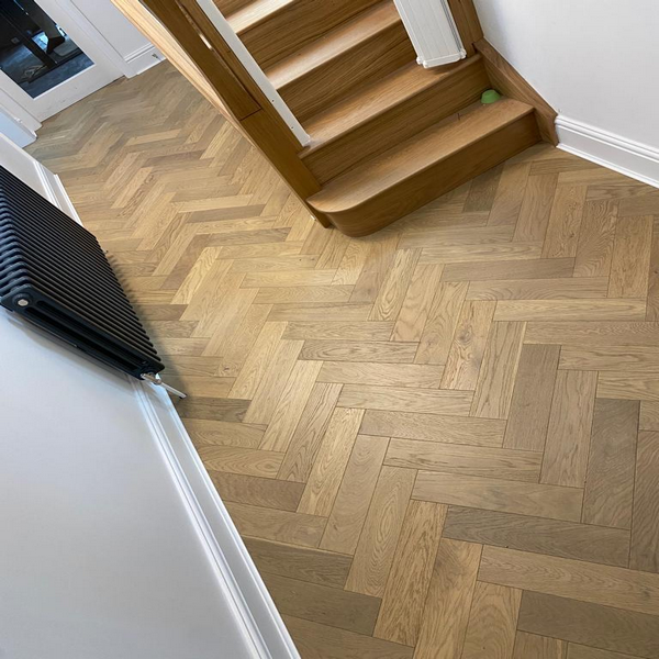 Herringbone-patterned wooden flooring in a hallway with a staircase and black radiator.