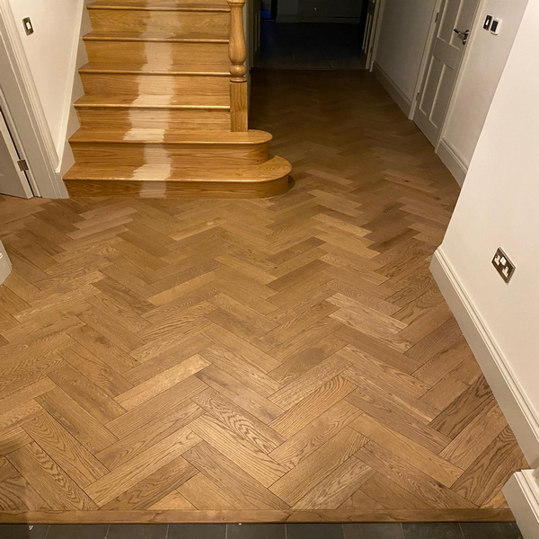 A hallway with herringbone-patterned wooden flooring and stairs, partially sanded on one side.