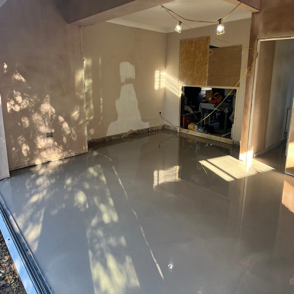 A room under renovation with freshly poured concrete floor, unfinished walls, exposed wiring, and lighting fixtures hanging from the ceiling.