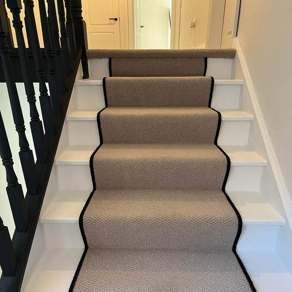 Staircase with a beige carpet runner bordered in black, white risers, and black railings.