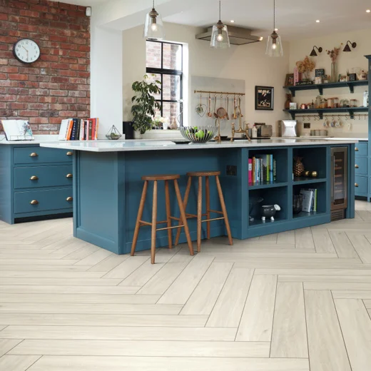 The modern kitchen features a herringbone wood flooring design, complemented by a blue island with bar stools. A brick wall and shelves adorned with decor add character, while pendant lights complete the look.